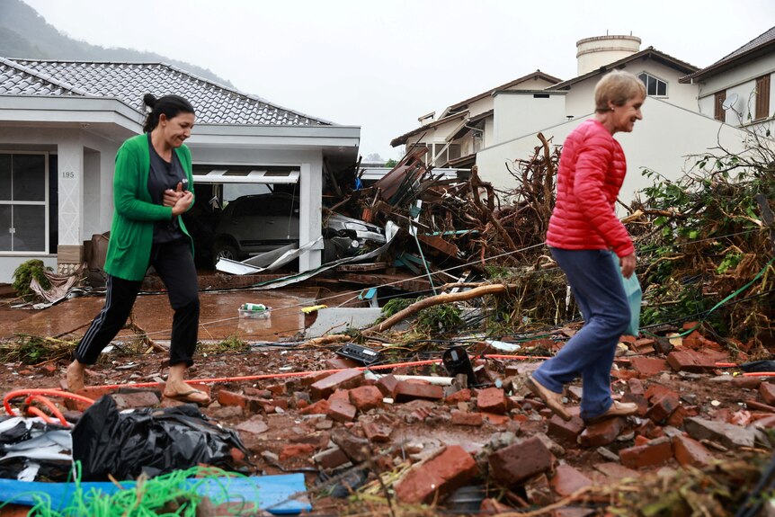 Two people walking over debris in front of houses. 