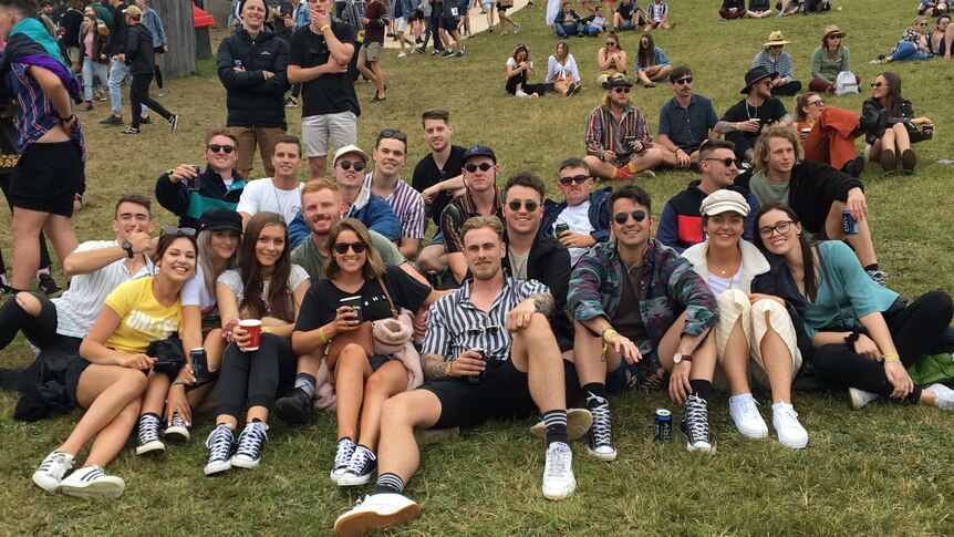 A group of young men and women sit on a grassy hill at a music festival