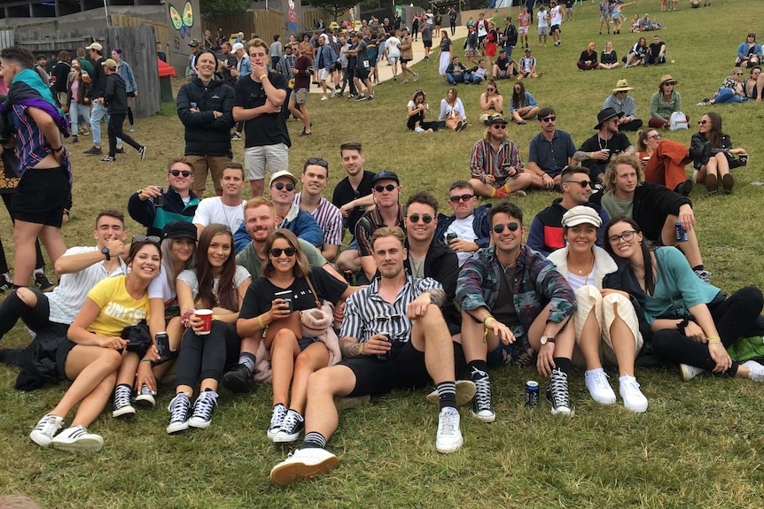 A group of young men and women sit on a grassy hill at a music festival