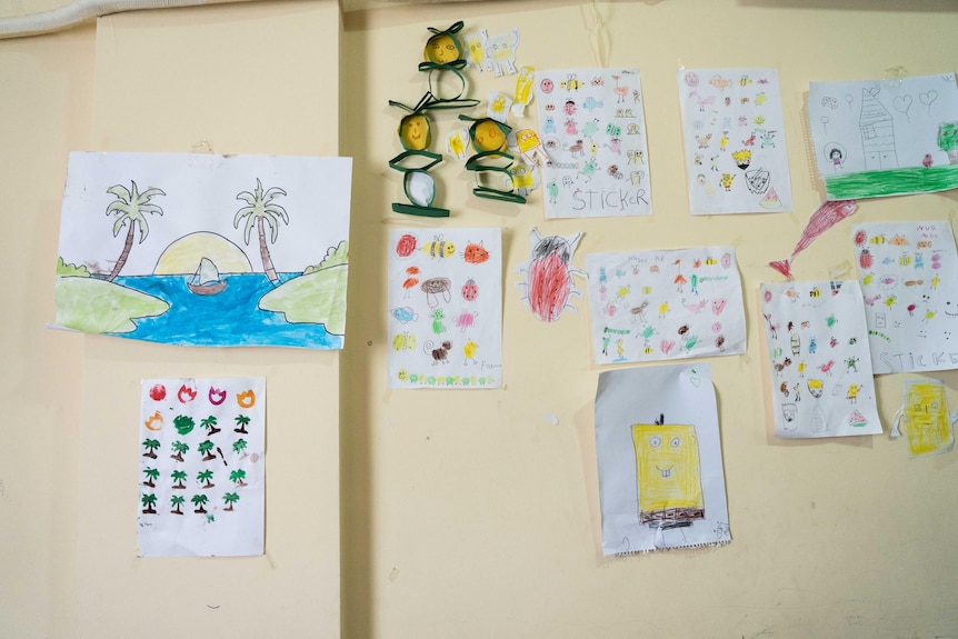 Drawings and paintaings created by Turkish and Syrian children are displayed on a yellow wall.