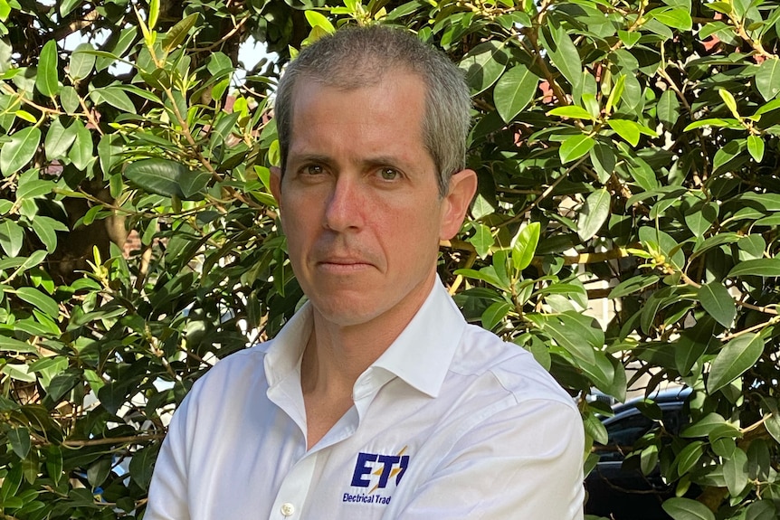 Man in a white collared shirt with a serious expression stands in front of a tree with his arms crossed.