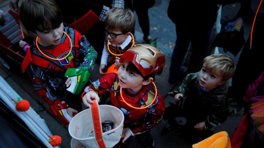 Four little boys in Halloween costumes wait at a door for candy, with one boy holding a bucket and looking up.