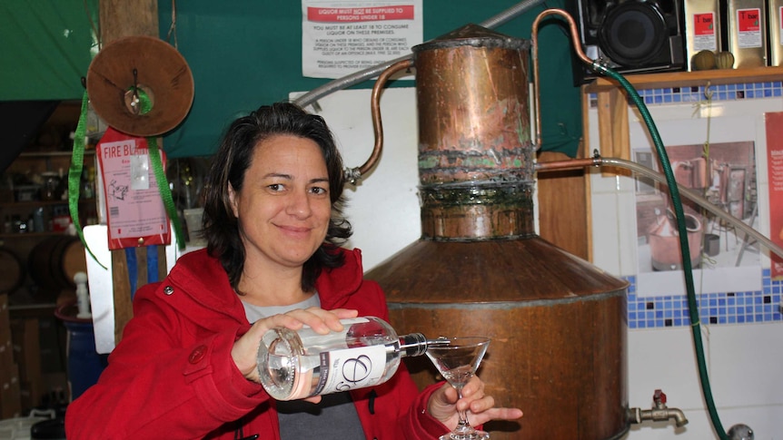 Sarah Lark pouring gin into a cocktail glass, standing in front of a copper still.