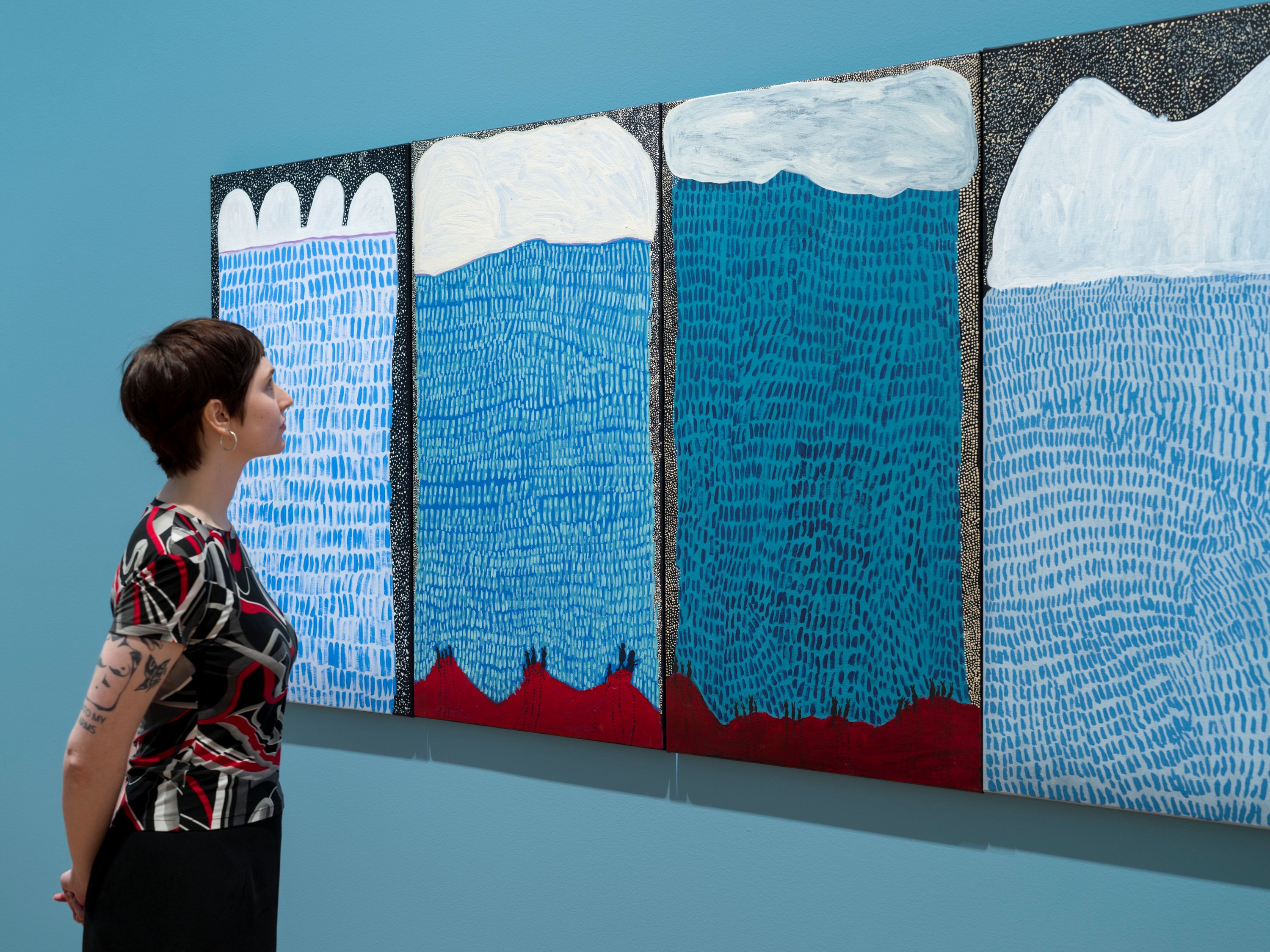 En una galería, una mujer se encuentra frente a cuatro paneles pintados que representan un cielo azul con patrones intrincados sobre un paisaje rojo.