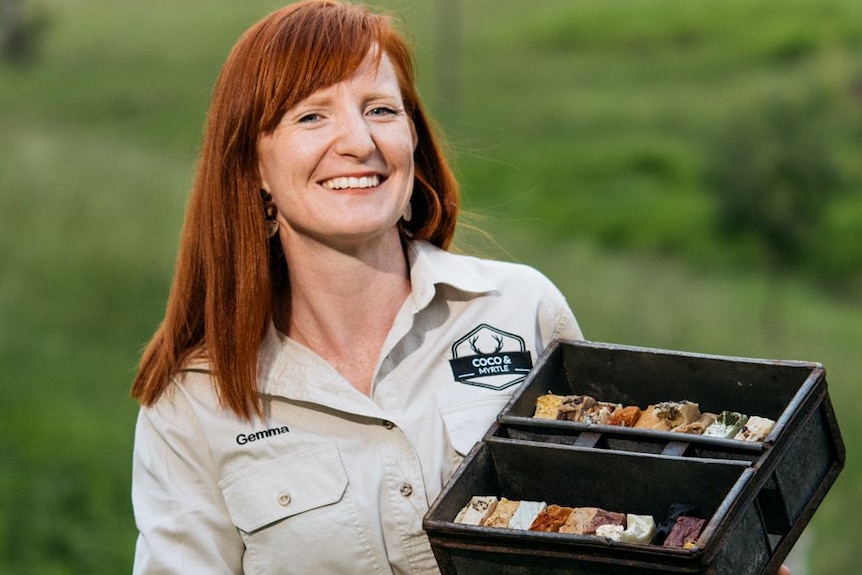 A red-haired smiling woman with bangs, khaki shirt with logo, holds box of soaps, blurred grassy area behind.