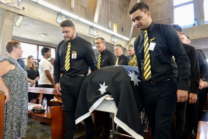 Male pallbearers dressed in black with yellow ties carrying a casket down the aisle of a church. 