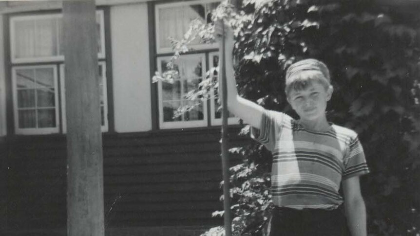 A black-and-white picture of a boy of about 10, in a striped T-shirt, holding a snake, outside a cottage.