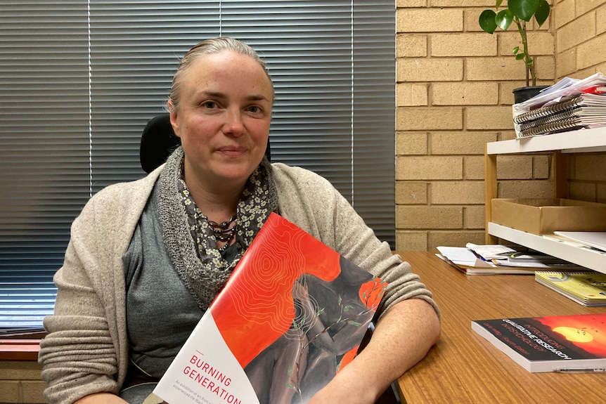 Rachael Fox sits in her office holding a booklet of the artwork in the Burning Generation.