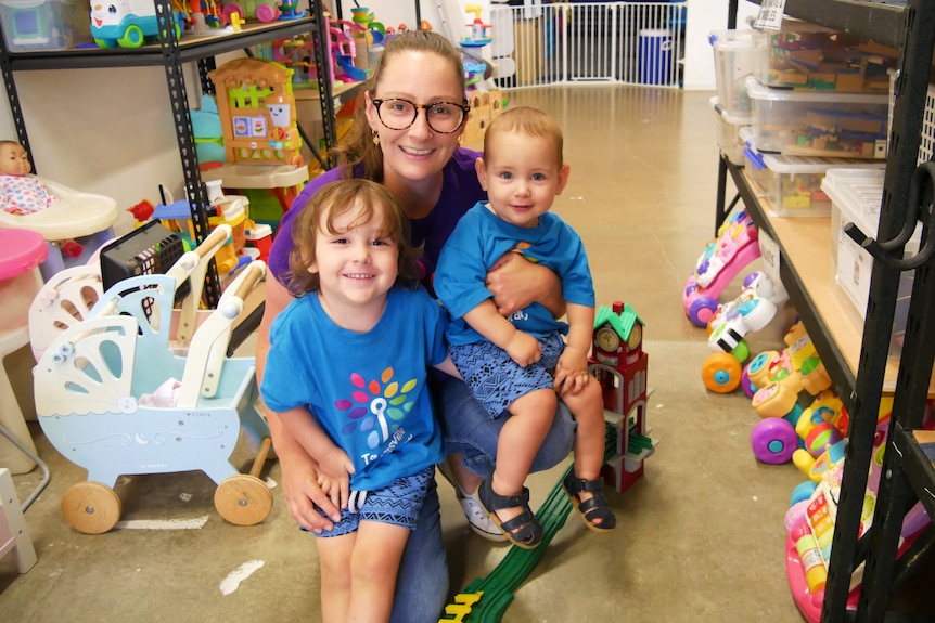 A woman with her arms around toy young boys surrounded by toys in a toy library