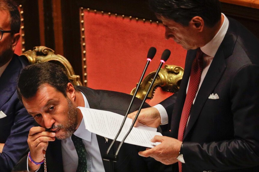 Man holds a rosary to his mouth and kisses it as prime minister stands next to him reading from a sheet of paper