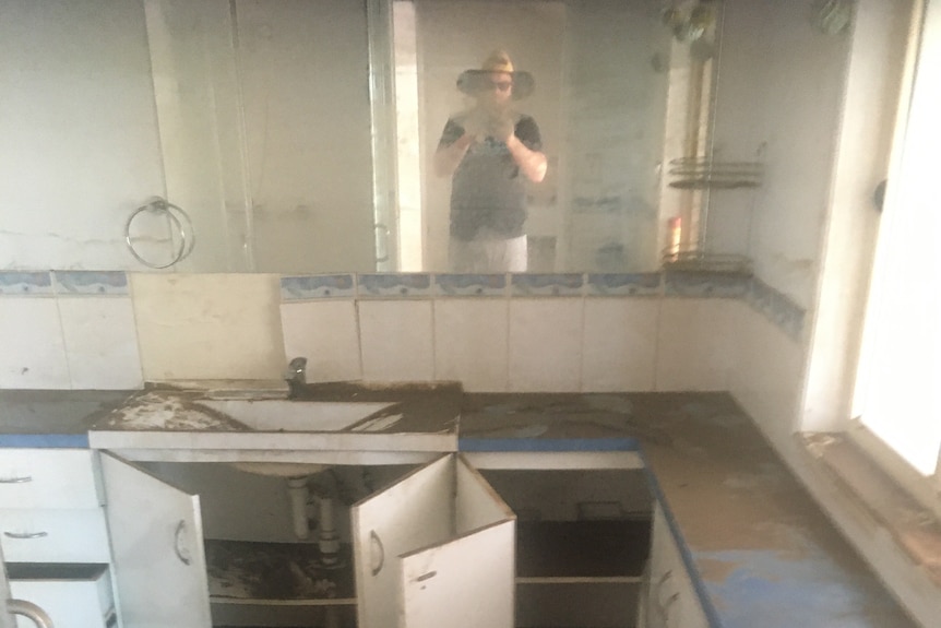 A man in a hat, who can be seen in a bathroom mirror, photographs the flood-damaged room.