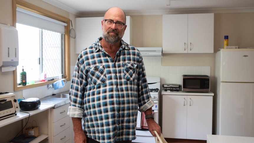 A man with a beard wearing a plaid shirt stands in a kitchen and looks at the camera.