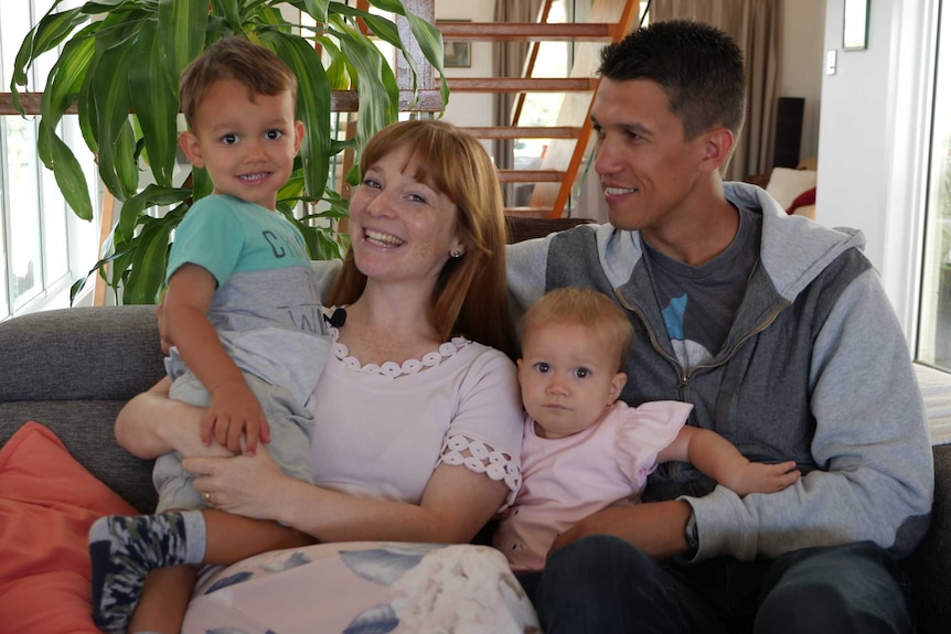 A mother and father sit on the lounge with a baby girl and young boy.