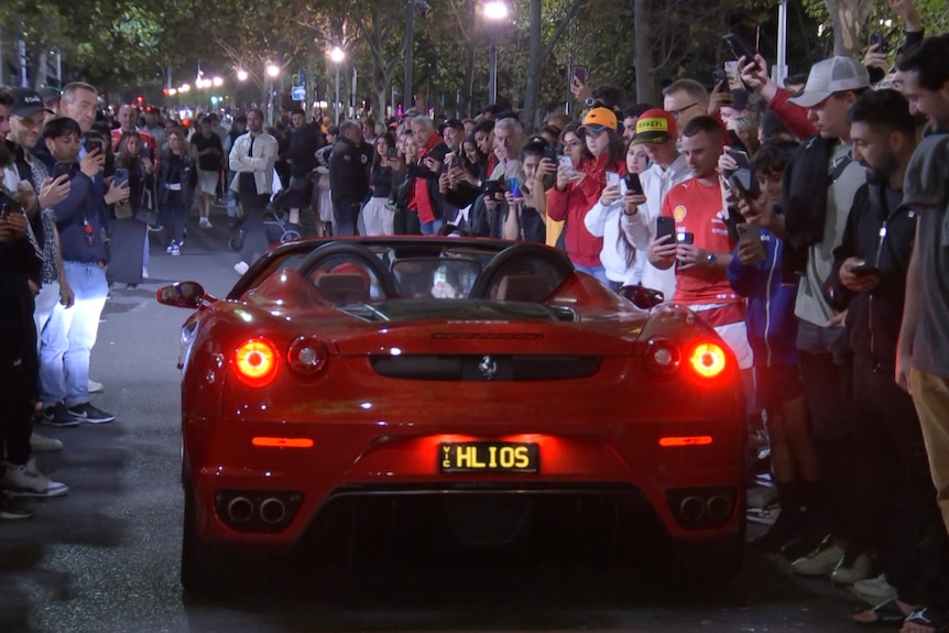 A red ferrari drives through a street lined with crowds of formula one fans.