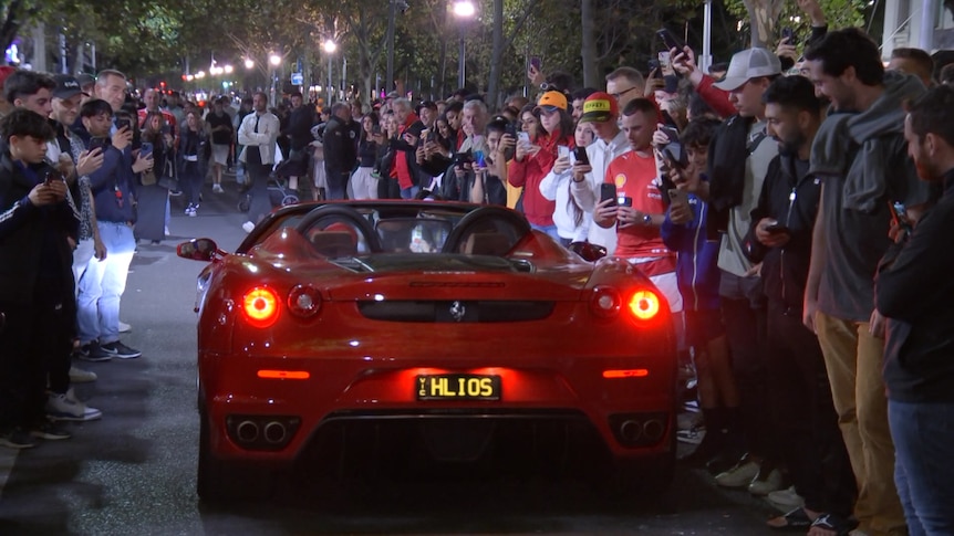 A red ferrari drives through a street lined with crowds of formula one fans.