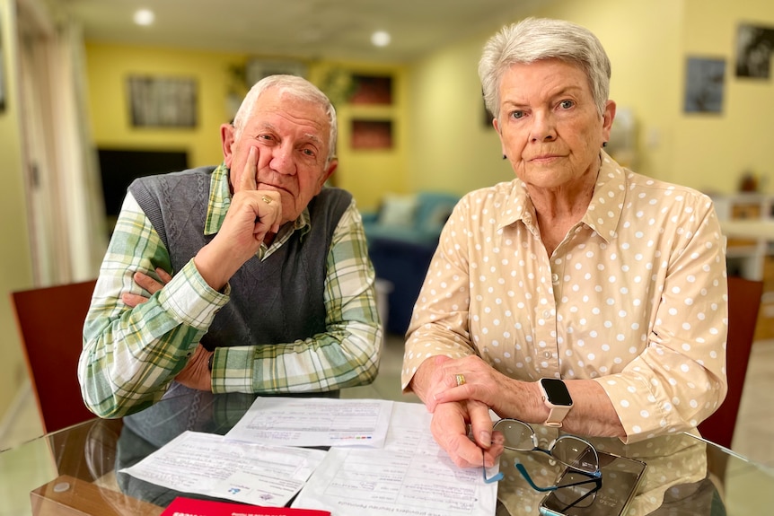 A couple with grey hair sit at a table, several documents in front of them, looking serious.