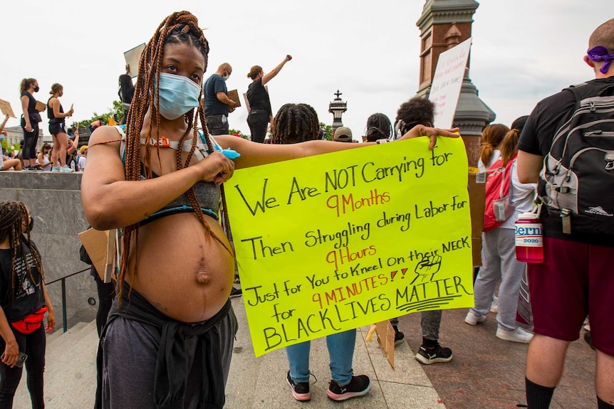 A pregnant woman holds a sign reading 'we are not carrying for 9 months then struggling during labour for 9 hours'