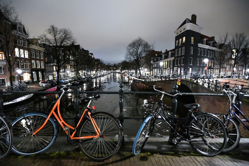 Bicycles lean up against the railing of a bridge over a canal in Amsterdam. Windows light up the dark cloudy evening sky
