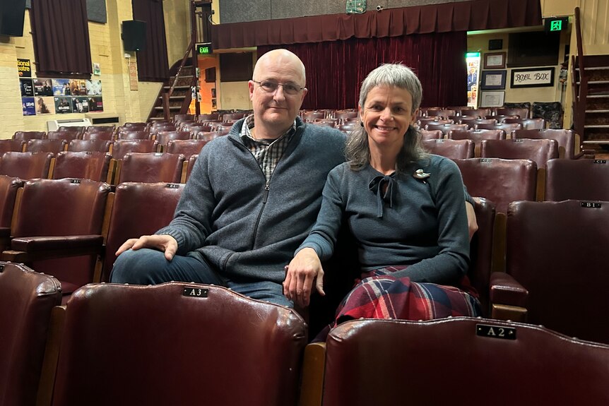 A man and a woman sit among rows of old-fashioned burgundy chairs in a hall.  
