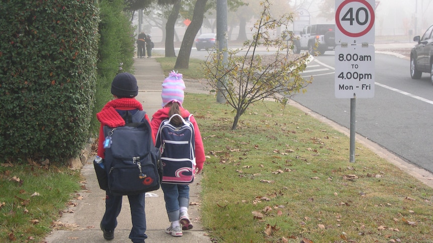 Two young children, wearing large backpacks, walk to school on a cold, foggy day. They are wearing beanies and scarves.