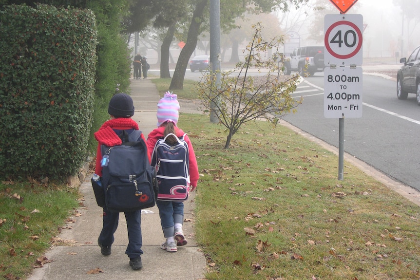 Two young children, wearing large backpacks, walk to school on a cold, foggy day. They are wearing beanies and scarves.