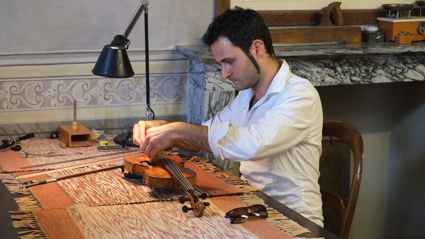 A man sits at a table cleaning and restoring a violin.