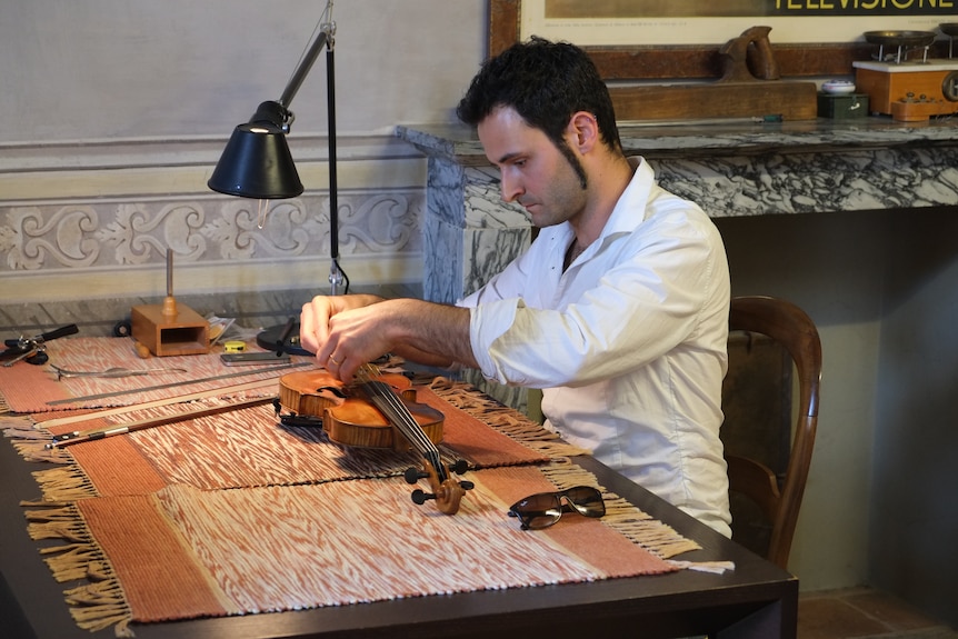 A man sits at a table cleaning and restoring a violin.