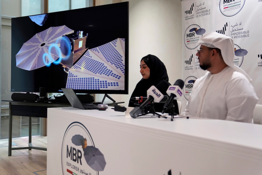 A man in white headdress and thawb and a woman in a black hijab sit next to a computer rendering of a spacecraft.
