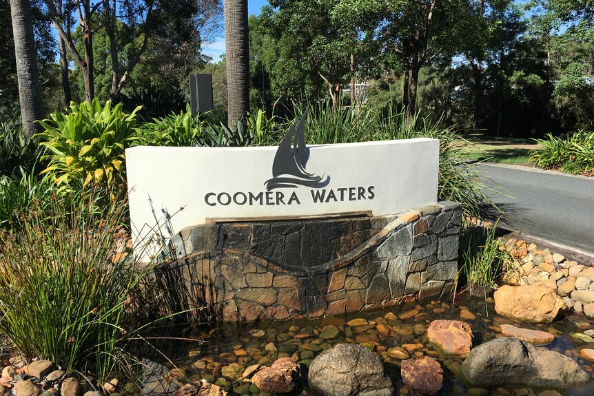 A roadside sign of Coomera Waters and a figure of a boat sailing on water surrounded by rocks and vegetation.