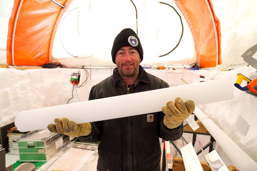 Dr Mark Curran wearing an Australian Antarctic Division beanie and thick yellow gloves, holds a cylindrical piece of ice