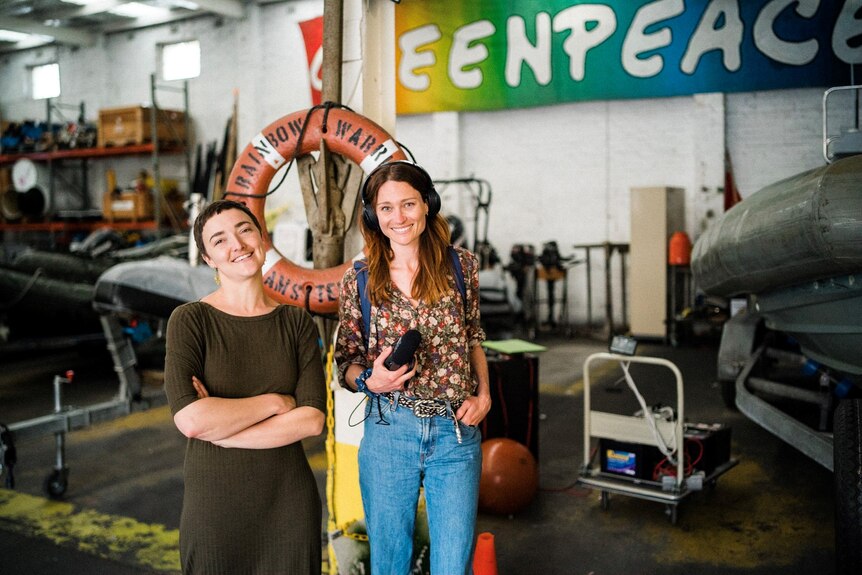 Two women, one with headphone and a recorder, pose for a photo together in the Greenpeace warehouse.
