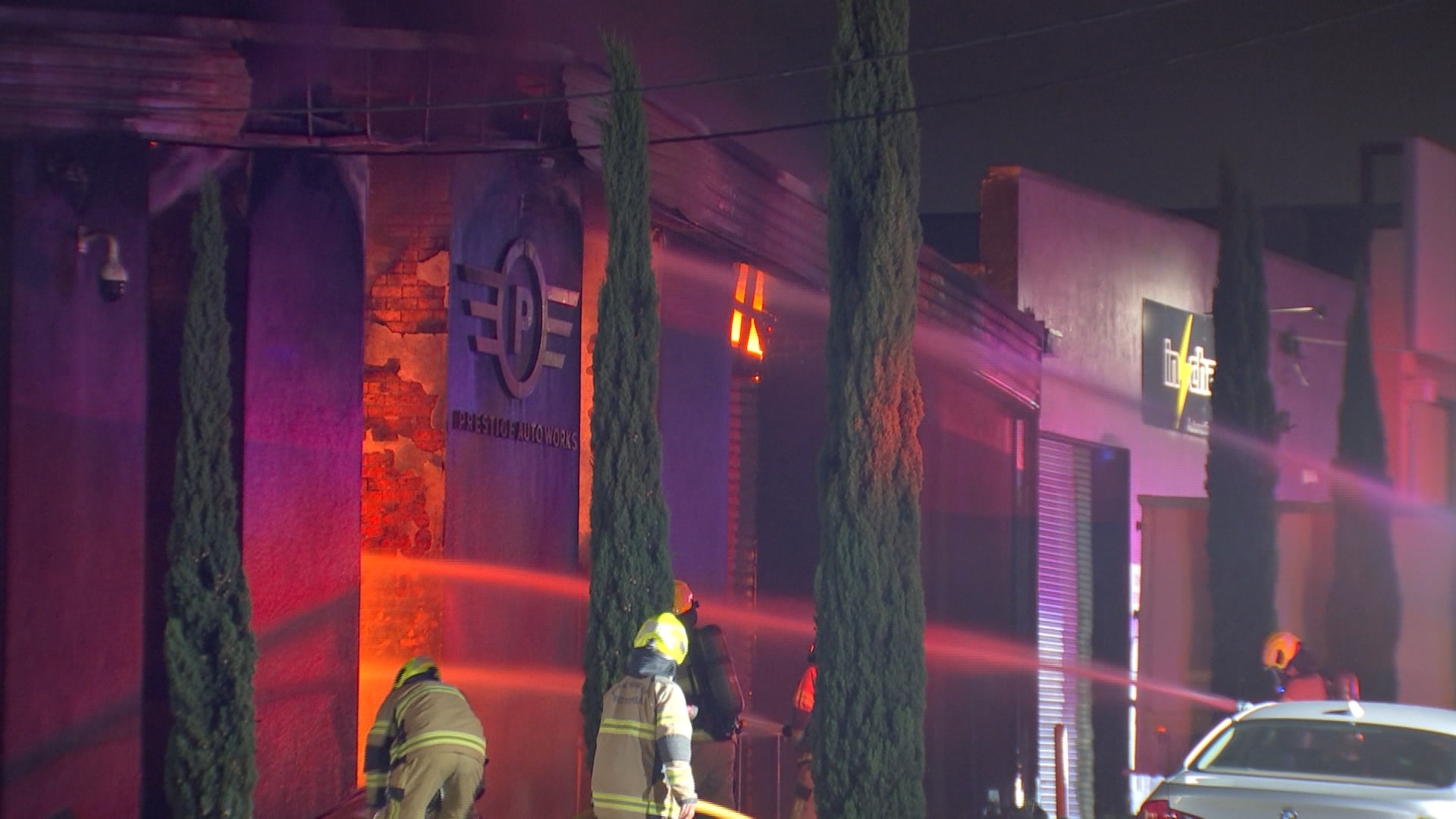Several firefighters point hoses at a building that has flames visible inside at night.