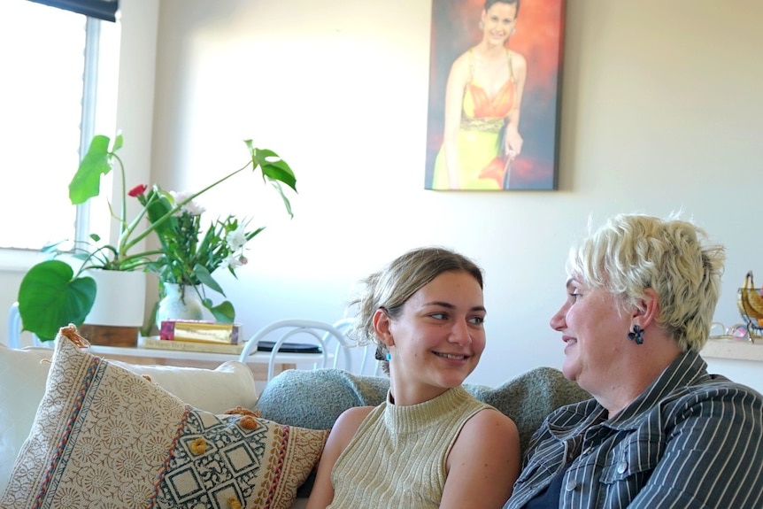 A mother and daughter cuddle up on a couch, looking at each other and smiling. On the wall behind them is a portrait 