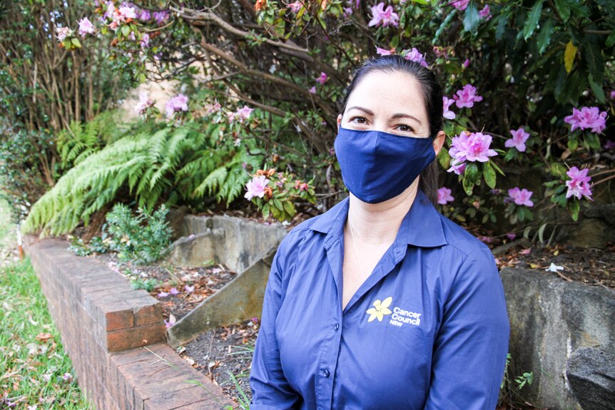 A woman in a dark-coloured shirt, wearing a mask and standing in a garden.