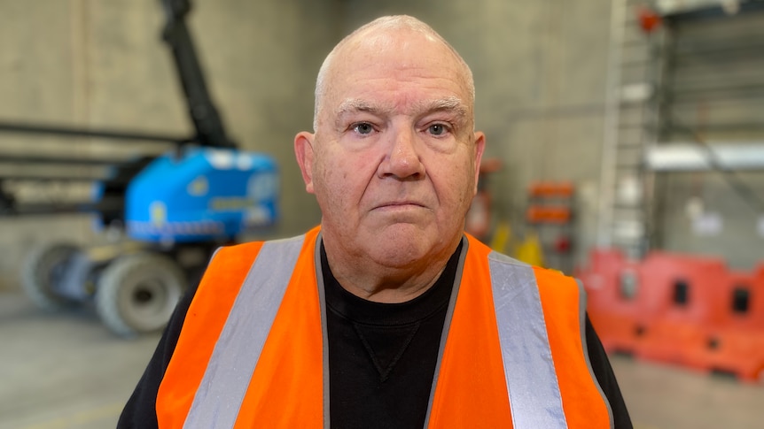 A bald man wearing a bright orange vests stares in the camera, behind him is heavy machinery in a warehouse.