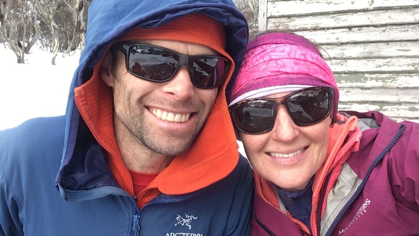 A man and woman wearing sunglasses and wearing snow gear smile for a selfie with snowy bush in the background.