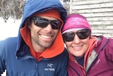 A man and woman wearing sunglasses and wearing snow gear smile for a selfie with snowy bush in the background.