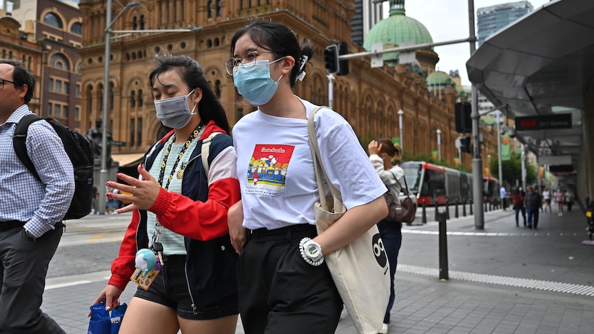 Two young women walk along a street wearing face masks. 