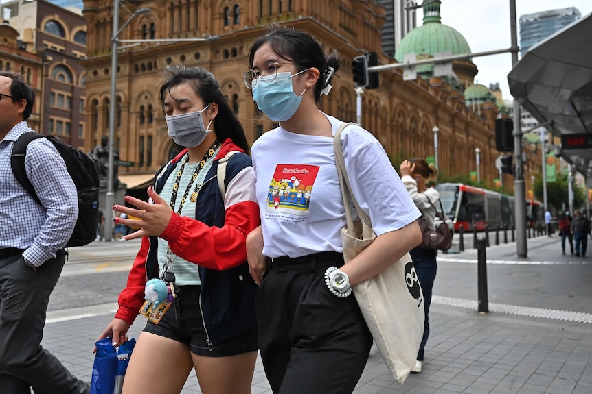 Two young women walk along a street wearing face masks. 