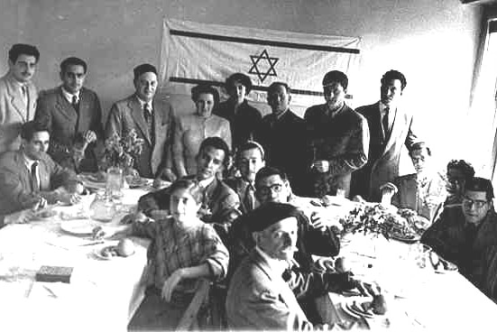 A dozen people at tables face the camera in front of an Israeli Flag