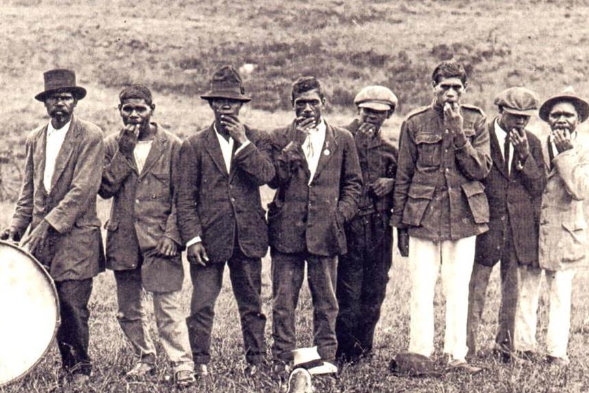 A black and white photo of a line of Indigenous men posing for a photo with a bass drum, standing in a park.