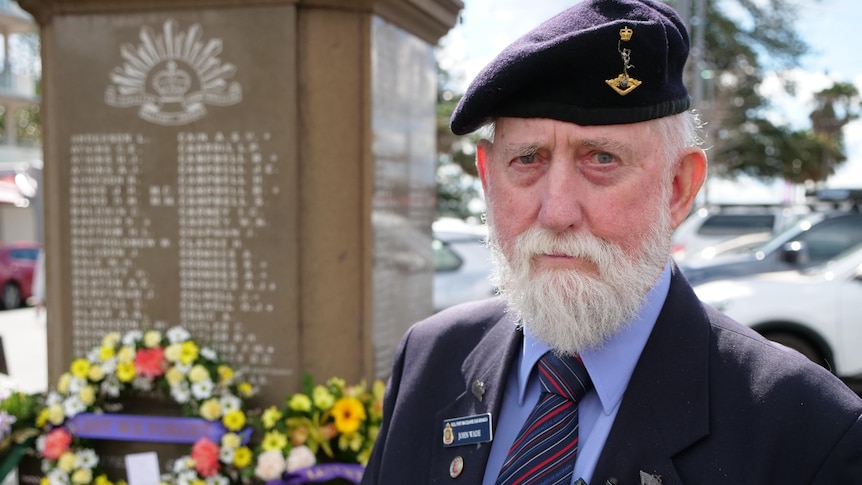 Mr Wade stands in front of a war memorial that is covered in flowers. 