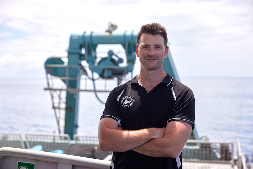 A man smiling with his arms folded with view of sea and diving equipment in background.