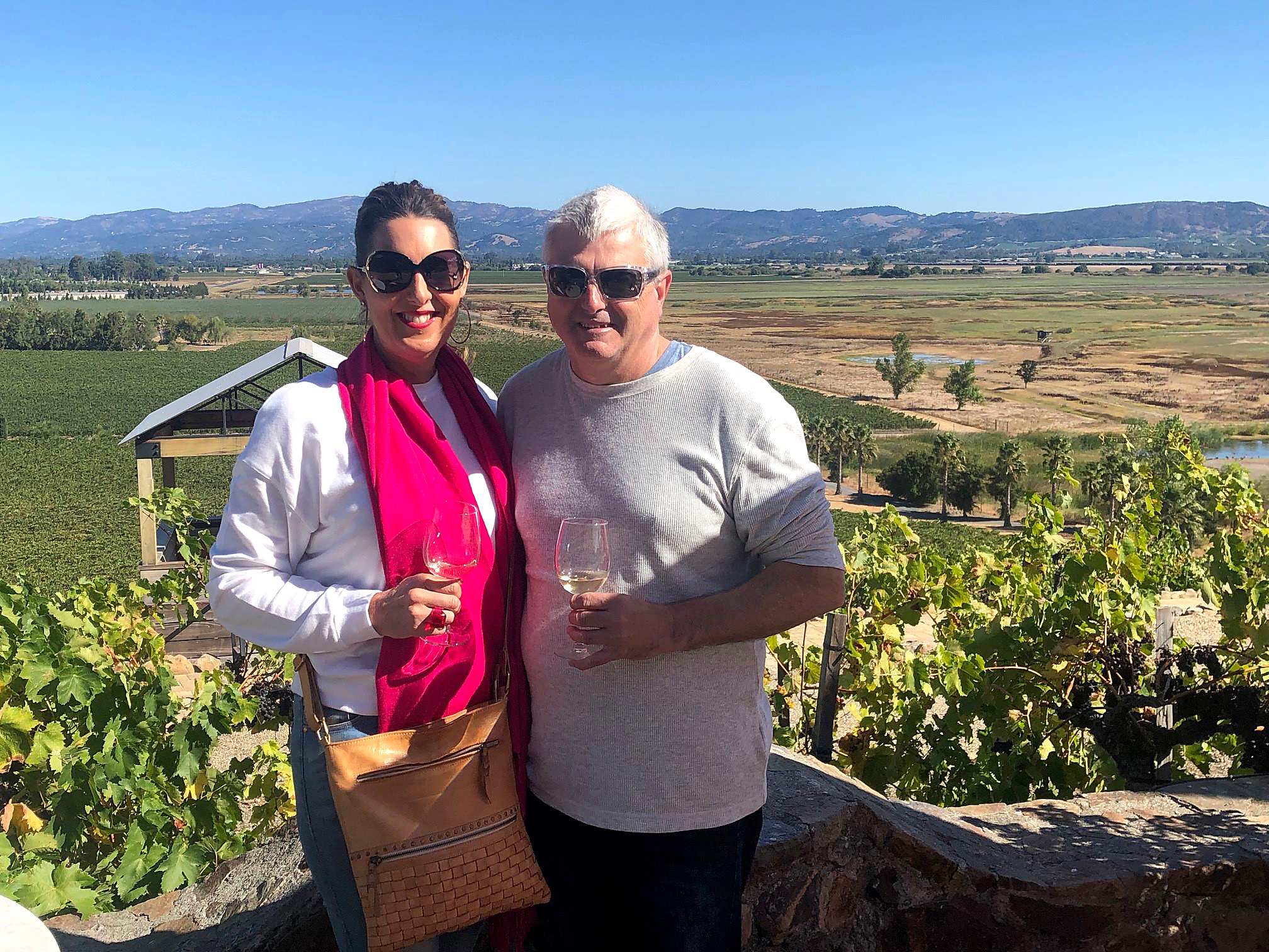 A woman and a man smile while holding wine glasses with a vineyard in the background.
