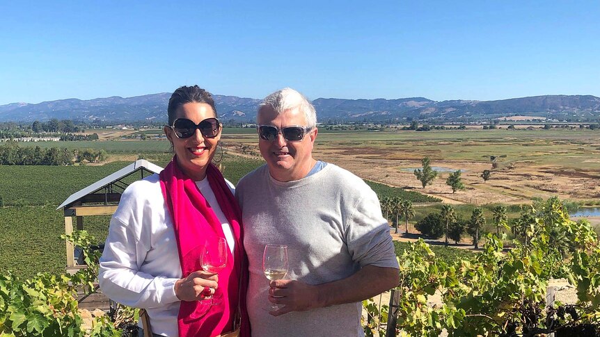 A woman and a man smile while holding wine glasses with a vineyard in the background.