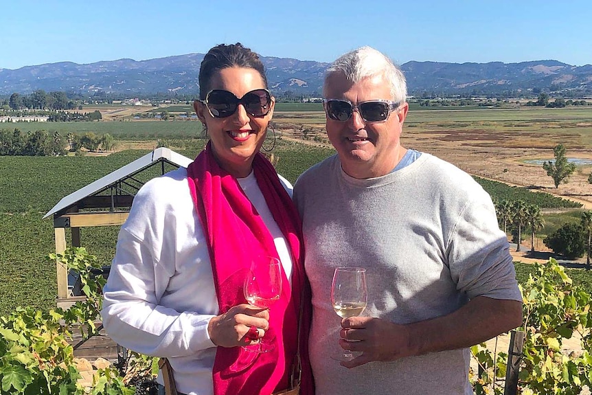 A woman and a man smile while holding wine glasses with a vineyard in the background.