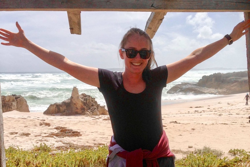 A woman with her arms up int he air and a smile on her face with the beach of Portland behind her