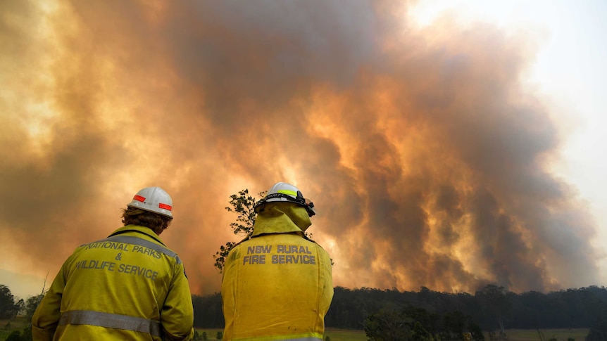 Two men in firefighter uniforms look towards a wall of smoke, backlight by fire, which fills the sky in a rural setting.