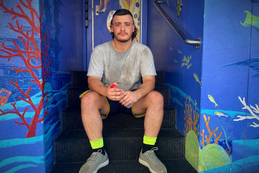 A man with a dirty grey shirt, neon yellow socks and a mullet sits on a stairwell with blue painted walls behind him. 
