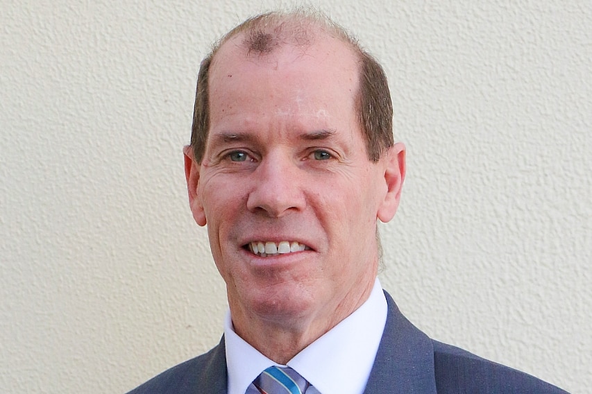 A balding man in a greying blue suit looks at the camera in a professional portrait